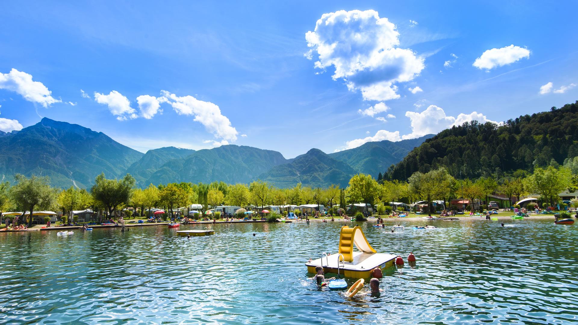 Lago alpino con montagne, campeggio e persone che nuotano.